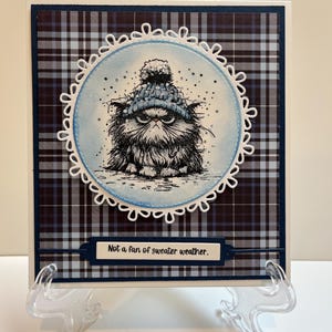 May include: Greeting card featuring a grumpy cat wearing a blue knit hat. The cat illustration is inside a white scalloped circle, set against a blue and white plaid background. Text reads, "Not a fan of sweater weather."