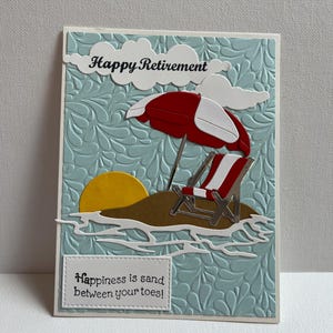 May include: A handmade retirement card with a beach theme. The card features a red and white striped beach chair, a red and white umbrella, and a yellow sun. The card reads "Happy Retirement" and "Happiness is sand between your toes!"