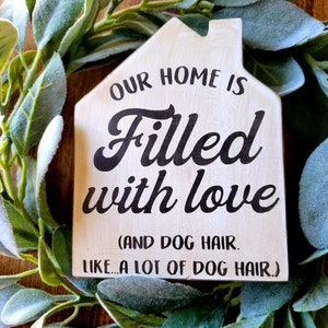 May include: A wooden house-shaped sign with the text "OUR HOME IS Filled with love (AND DOG HAIR. LIKE...A LOT OF DOG HAIR.)" in black lettering. The sign is surrounded by a green and gray leafy wreath.