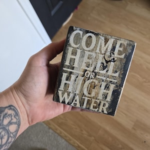 Puede incluir: Un letrero rectangular de madera con el texto "COME HELL OR HIGH WATER" en letras blancas. El letrero tiene un aspecto envejecido y rústico con una combinación de colores marrón oscuro y beige.