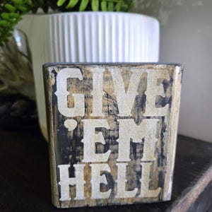 May include: A rustic wooden block with the words "GIVE 'EM HELL" in a distressed white font. The block has a dark, weathered appearance with visible wood grain and a touch of gold. The background includes a white vase and greenery.