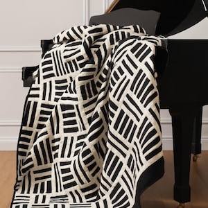 Può includere: Una trapunta a motivi geometrici in bianco e nero drappeggiata su un pianoforte a coda nero. La trapunta presenta un motivo ripetuto di linee nere su uno sfondo bianco sporco. Il pianoforte è appoggiato su un pavimento in legno.