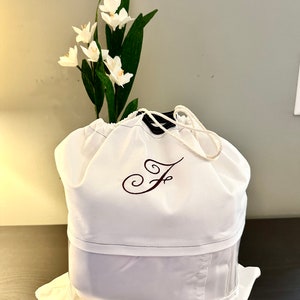 May include: White drawstring bag with a clear window, featuring a burgundy embroidered letter "F" on the front. The bag is sitting on a dark brown surface with white flowers in the background.