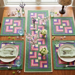 Peut inclure: Une table à manger en bois avec un ensemble de sets de table et de chemins de table matelassés verts, roses et violets. Chaque couvert comprend une assiette blanche, une serviette verte et des couverts. Des compositions florales et de la verrerie complètent la table.