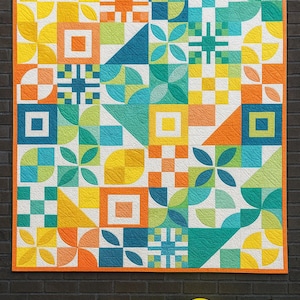 Puede incluir: Un edredón colorido con patrones geométricos en tonos de amarillo, naranja, turquesa y azul. El edredón tiene un borde naranja y cuelga contra una pared de ladrillos oscuros. Globos de varios colores en primer plano.