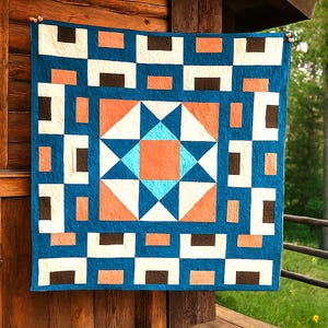 May include: A handmade quilt with a geometric design. The quilt features a teal background with white, brown, and orange shapes. The central design is a starburst pattern in shades of teal, orange, and white. The quilt is hanging on a wooden structure.