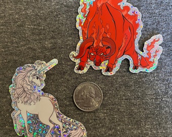 May include: Two holographic stickers, one of a red bull with flames and the other of a white unicorn. Both stickers have a glittery finish.