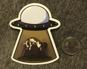 May include: A sticker featuring a cartoon UFO with a cow being abducted by a tractor beam. The UFO is black and gray with green lights. The cow is brown and white. A quarter is next to the sticker for size comparison.