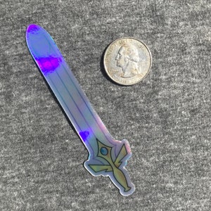 Op de afbeelding: Een holografische sticker van een zwaard met een blauwe en paarse iriserende afwerking. Het zwaard heeft een gouden handvat en een blauwe edelsteen in het midden. De sticker is ongeveer zo groot als een Nederlandse euro.