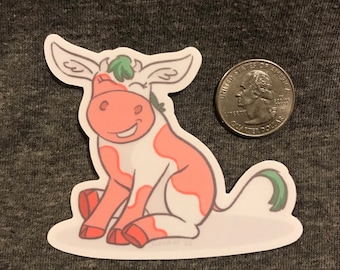 May include: A pink and white cow sticker with green accents. The cow is sitting down with its tail curled up behind it. The sticker is on a gray background.