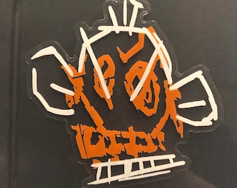May include: A white sticker with a brown outline of a cartoon face with a large nose and eyes. The face is drawn with simple lines and has a whimsical, childlike style.