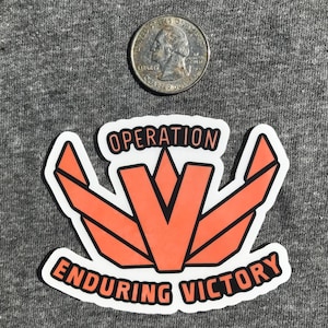 May include: A white sticker with a red and black geometric design. The sticker says "Operation Enduring Victory" and features a stylized V shape.