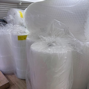 May include: Large cylindrical rolls of white bubble cushioning wrap protected by clear plastic film. Each roll features a yellow rectangular label with black text, indicating the product is designed for protective packaging and shipping needs.