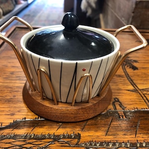 May include: A vintage serving bowl with a black lid and a white body with black vertical stripes. The bowl sits in a gold-colored metal frame on a wooden base. A small red dot is visible on the bowl's side.