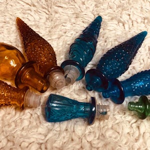 Peut inclure: Une collection de bouchons de bouteilles décoratifs en verre de différentes couleurs, dont ambre, marron, bleu sarcelle et vert. Les bouchons ont des motifs coniques texturés et sont disposés sur une surface blanche et douce.