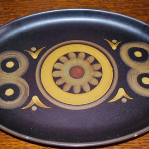 May include: Dark brown oval ceramic platter with a decorative pattern. The centre has a yellow flower with a red centre, surrounded by concentric circles and stylized shapes in yellow and brown.