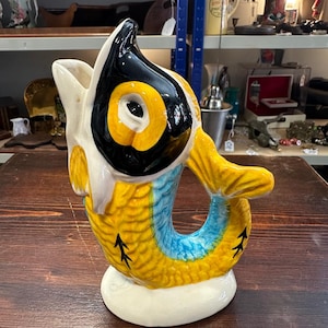 May include: A ceramic fish-shaped pitcher with a white base. The pitcher features a yellow, blue, and black color scheme. The fish has a large eye and a handle that forms the tail. The pitcher is sitting on a wooden surface.
