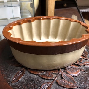 May include: A two-tone ceramic pudding mold with a scalloped rim. The mold features a light beige interior and a brown exterior band and rim. The mold is sitting on a dark wooden surface with a leaf design.
