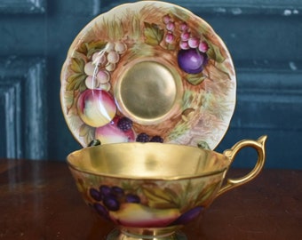 May include: A vintage teacup and saucer set with a gold rim and a fruit motif. The cup and saucer are decorated with a colorful painting of fruit, including grapes, plums, and apples.