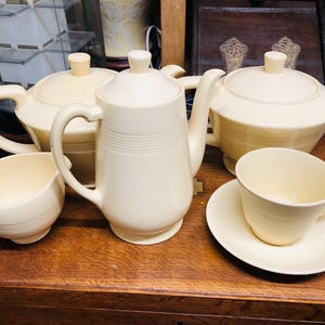 Könnte beinhalten: Ein cremefarbenes Teeservice, bestehend aus einer Teekanne, einer Kaffeekanne, einem Sahnekännchen, einer Zuckerdose, einer Tasse und einer Untertasse, auf einer Holzoberfläche angeordnet. Das Set hat ein klassisches Design mit glatter Oberfläche und dezenten Details.