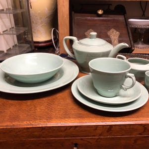 May include: A full set of pale green tableware, including a teapot, teacup, saucer, bowl, creamer, and small cup. The items are displayed on a wooden surface, presenting a classic, coordinated dining set. The set is suitable for serving tea or coffee.