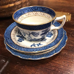 Puede incluir: Un juego de taza y platillo azul y blanco con ribete dorado. La taza y el platillo presentan un patrón detallado de árboles y pájaros. El juego está colocado sobre una superficie de madera, lo que sugiere una estética vintage o antigua.