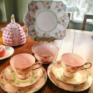 Könnte beinhalten: Ein Vintage-Tee-Set mit rosafarbenem und floralem Design. Das Set enthält eine Teekanne, eine Zuckerdose, zwei Teetassen und zwei Untertassen. Die Teekanne ist in Form einer Frau in einem rosafarbenen Kleid. Die Teetassen und Untertassen haben ein zartes Blumenmuster.