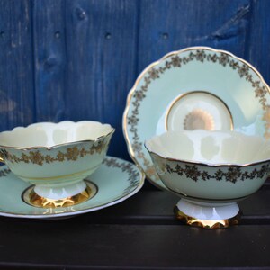 May include: Two delicate teacups and saucers with a light green and gold floral design. The cups and saucers have a scalloped edge and a gold trim.