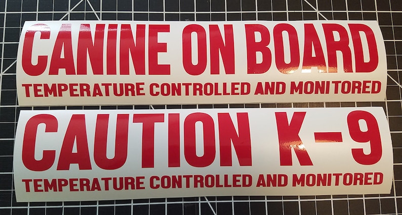 Op de afbeelding: Twee rechthoekige witte borden met rode tekst. Het bovenste bord heeft de tekst "CANINE ON BOARD TEMPERATURE CONTROLLED AND MONITORED". Het onderste bord heeft de tekst "CAUTION K-9 TEMPERATURE CONTROLLED AND MONITORED".