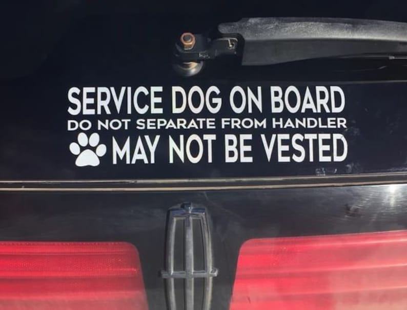 May include: A black car bumper with a white sticker that reads "SERVICE DOG ON BOARD." Below the text are the words "DO NOT SEPARATE FROM HANDLER" and "MAY NOT BE VESTED." A paw print graphic is also included.