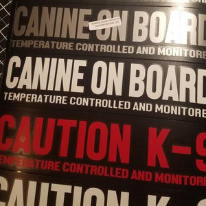 Op de afbeelding: Drie zwarte en zilveren reflecterende stickers met witte tekst die "CANINE ON BOARD TEMPERATURE CONTROLLED AND MONITORED" luidt. De derde sticker is rood en zwart en luidt "CAUTION K-9 TEMPERATURE CONTROLLED AND MONITORED".