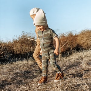 May include: Two children wearing beanies and casual clothing walk through a field of dry grass. The child in the front is wearing a camouflage print jumpsuit and brown boots.