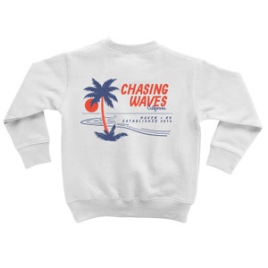 Puede incluir: Sudadera blanca con un gráfico azul y rojo de una palmera, sol y olas. El texto "Chasing Waves California Haven -  KO Established 2014" está impreso en la sudadera.