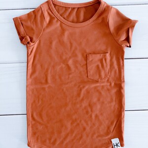 May include: A rust-colored short-sleeved t-shirt with a chest pocket. The shirt has a rounded hem and rolled-up sleeves. A small white tag with black text is visible on the lower right side of the shirt.