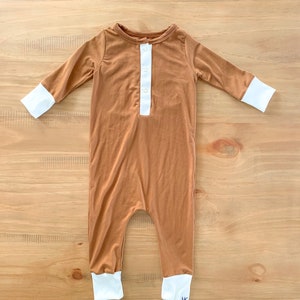 May include: A brown baby onesie with long sleeves and white cuffs. The onesie has a white button-up placket and white cuffs on the sleeves and legs. The onesie is made of a soft, comfortable material.