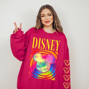 May include: A pink sweatshirt with a tie-dye Mickey Mouse graphic and the word "DISNEY" in yellow letters. The sleeves have a repeating pattern of yellow Mickey Mouse heads.