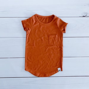 May include: A burnt orange t-shirt with a small pocket on the chest. The short-sleeved shirt has a rounded hem and a small white tag with black text near the bottom. The shirt is made of a soft material.