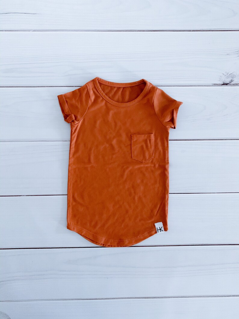 May include: A burnt orange t-shirt with a small pocket on the chest. The short-sleeved shirt has a rounded hem and a small white tag with black text near the bottom. The shirt is made of a soft material.