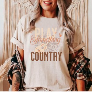 May include: A beige t-shirt with the text "Play Something Country" in brown and orange lettering. A stylized guitar with stars and birds is featured above the text.