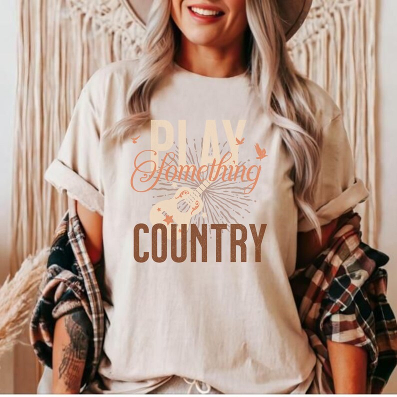 May include: A beige t-shirt with the text "Play Something Country" in brown and orange lettering. A stylized guitar with stars and birds is featured above the text.