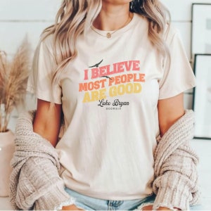 May include: A white t-shirt with a vintage-style graphic that reads "I believe most people are good" with the text "Luke Bryan Georgia" below. The graphic is in shades of red, orange, and yellow.