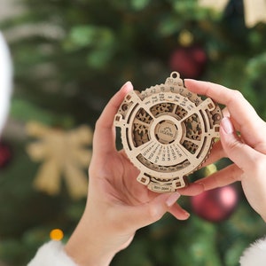 Op de afbeelding: Een houten eeuwigdurende kalender ornament met ingewikkelde tandwielen en een cirkelvormig ontwerp. De kalender toont de maanden van het jaar, de dagen van de week en de nummers 1-31. De ornament is gelabeld "Ugears" en "2021-2044".