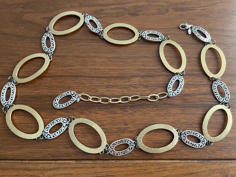 Puede incluir: Un collar dorado y plateado con eslabones ovalados. El collar presenta eslabones ovalados dorados y plateados alternados, con un cierre de mosquet&oacute;n. El collar se muestra sobre una superficie de madera.