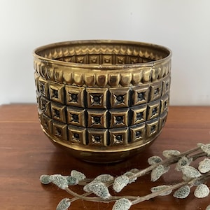 May include: A round brass planter featuring a textured exterior with a repeating grid pattern of raised squares and dark recessed centres. The upper rim is decorated with a scalloped border, and the metal has a warm, aged golden patina.