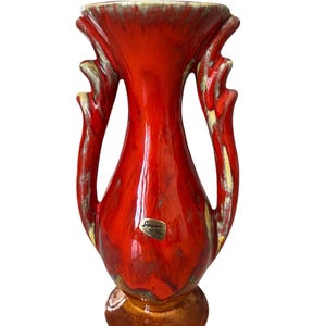 May include: A vibrant red ceramic vase with a flared top and two curved handles. The vase has a brown base and a label. The design features a blend of red, brown, and yellow hues, creating a unique decorative piece.