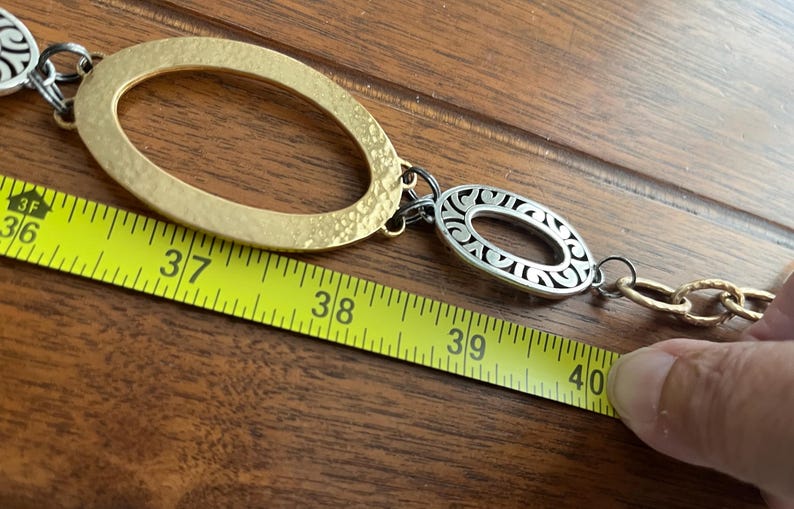 Puede incluir: Un cintur&oacute;n de metal dorado y plateado con eslabones ovalados y de cadena. El cintur&oacute;n se mide con una cinta m&eacute;trica amarilla, que muestra una longitud de aproximadamente 102 cm.