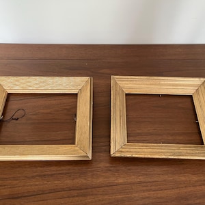 May include: Two square picture frames with a metallic gold finish. The left frame features a textured, wavy pattern on its surface, while the right frame displays a series of parallel, linear grooves. Both frames include a black hanging wire.