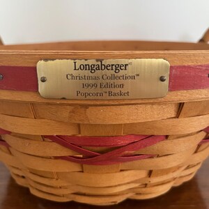 May include: A woven wooden basket featuring natural tan and red horizontal splints. A brass-coloured metal plate is attached to the front with the text: Longaberger Christmas Collection 1999 Edition Popcorn Basket.