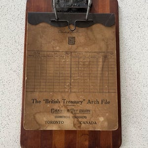 May include: A vintage "British Treasury" Arch File. The wooden clipboard has a metal clip and a paper insert with a grid and text. The text includes "Grand & Toy Limited, Commercial Stationers, Toronto, Canada."