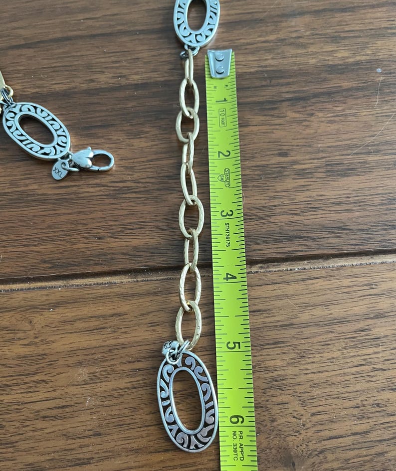 Puede incluir: Cintur&oacute;n de cadena plateada y dorada con eslabones decorativos ovalados. El cintur&oacute;n se mide con una cinta m&eacute;trica amarilla, mostrando una longitud de aproximadamente 15 cm. El cintur&oacute;n tiene un cierre de mosquet&oacute;n.